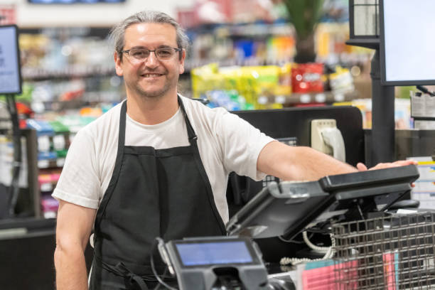 スーパーマーケットレジカウンターで働く成熟した男 - supermarket sales clerk grocer apron ストックフォトと画像