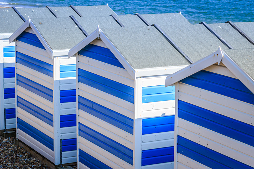 Row of three beach huts, all with blue doors. A mound of sand has blown up against them. Bright blue sky on a sunny day