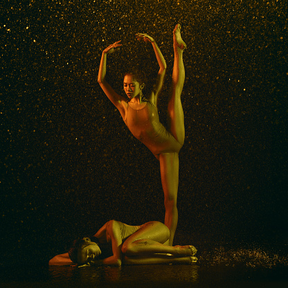 Sleep. Two young female ballet dancers under water drops and spray. Caucasian and asian models dancing together in neon lights. Ballet and contemporary choreography concept. Creative art photo.