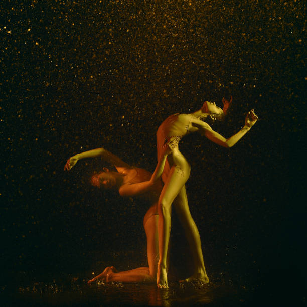 Flexibility. Two young female ballet dancers under water drops and spray. Caucasian and asian models dancing together in neon lights. Ballet and contemporary choreography concept. Creative art photo.