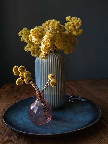 still life of vase with yellow flowers on a blue plate, wooden table against blue-green wall