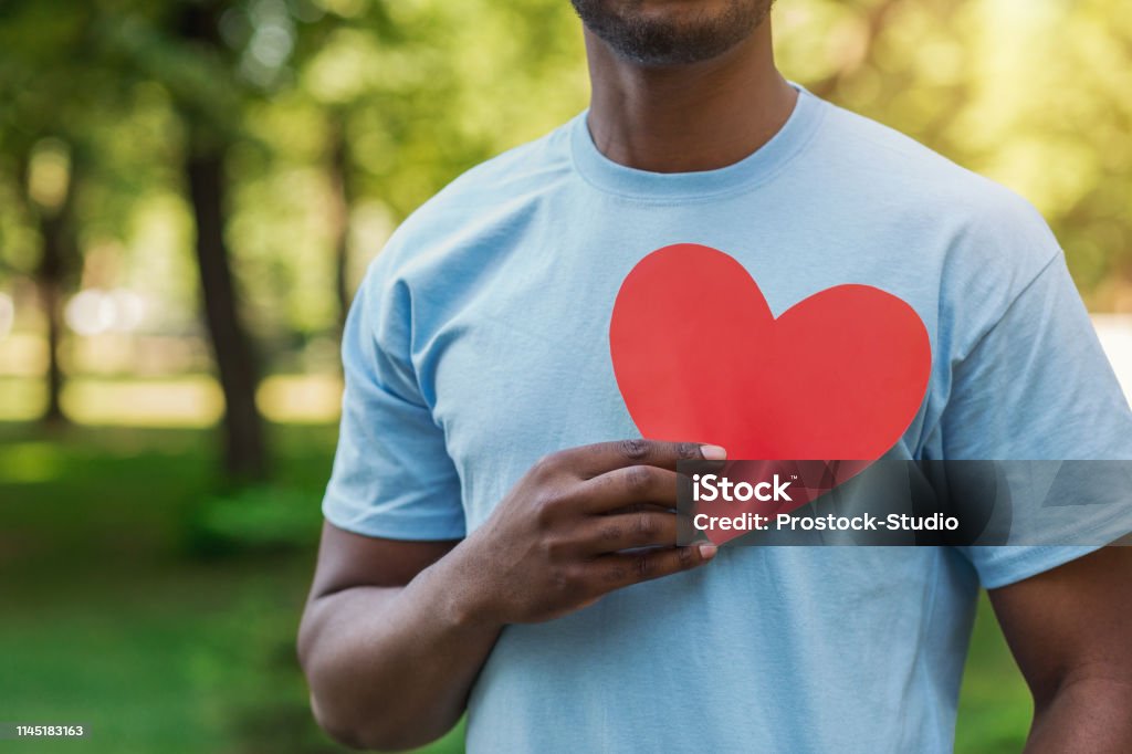 Black man holding red heart on his chest Love, volunteering and charity concept, copy space. Young black man holding red heart on his chest. Heart Shape Stock Photo