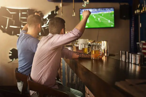 Two friends watching football game and drinking beer at the pub