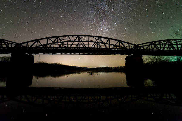 istock Illuminated metal bridge on concrete supports reflected in water on dark starry sky with Milky Way constellation background. Night photography concept. 1145195711