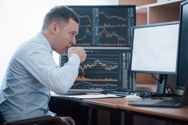 istock Drinks some coffee to stay concentrated. Man working online in the office with multiple computer screens in index charts 1145204281