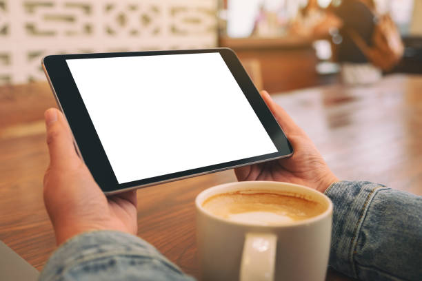 istock Mockup image of hands holding black tablet pc with blank white screen horizontally with coffee cup on wooden table 1145207360