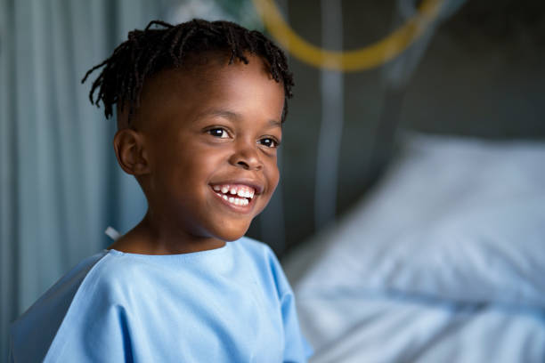 Smiling boy looking away at children's hospital Smiling boy looking away sitting on bed at ward. Cute male patient is in blue gown. He is at hospital. sick child hospital bed stock pictures, royalty-free photos & images