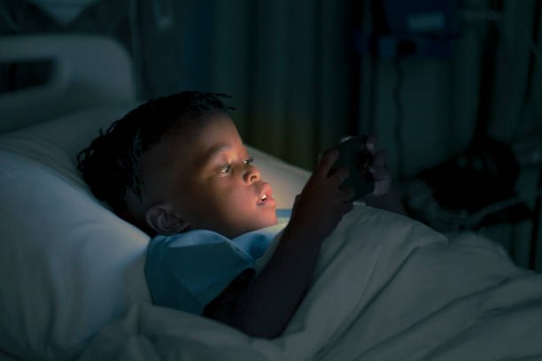 Ill boy using smart phone lying on bed in dark Ill male patient using mobile phone in dark at ward. Cute boy is lying on hospital bed at night. He is at children's hospital. night hospital bed stock pictures, royalty-free photos & images