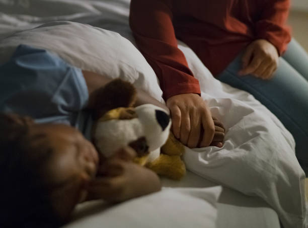 Mother holding ill son's hand sleeping on bed Midsection of woman holding son's hand sleeping on hospital bed. Ill boy is with stuffed toy relaxing by mother at ward. They are at children's hospital. sick child hospital bed stock pictures, royalty-free photos & images