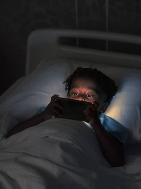 Ill boy using smart phone on bed in dark High angle view of ill male patient using mobile phone in dark. Cute boy is lying on hospital bed at ward. He is in children's hospital at night. night hospital bed stock pictures, royalty-free photos & images