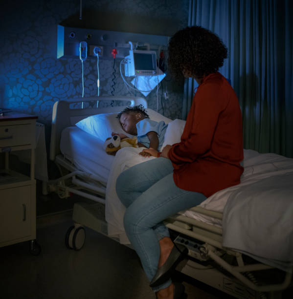 Young mother sitting by ill son sleeping on bed Young woman sitting by ill son sleeping on hospital bed. Boy is relaxing by mother in ward. They are at children's hospital. night hospital bed stock pictures, royalty-free photos & images