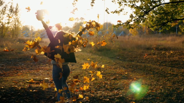 Young woman raising her hands and spinning around scattering autumn leaves. Happy girl showing joyful emotions with sunset at background. Lady enjoying beautiful autumn environment. Slow motion Close up