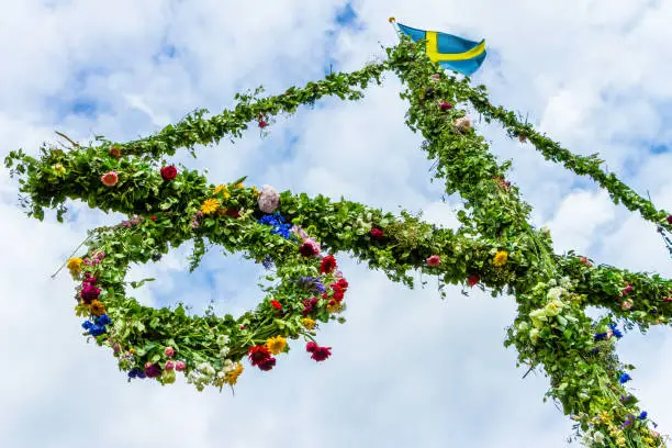 A pole and flag against the blue sky and white clouds. A maypole decorated, covered in flowers and leaves. Pole for celebrating midsummer. holiday. Midsummer traditional Swedish symbol.