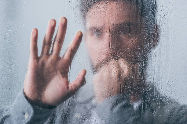 istock upset man covering mouth with hand and touching window with raindrops 1145223754