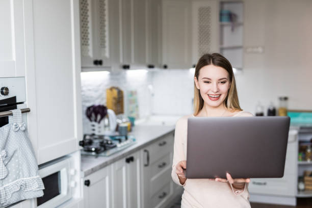 istock Smiling young woman using laptop in the kitchen at home 1145233711