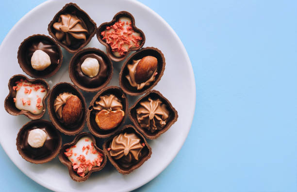 hermosos chocolates de diferentes formas y rellenos se encuentran en una placa blanca contra el papel de fondo azul. copiar espacio, vista superior. - peanut bowl nut circle fotografías e imágenes de stock