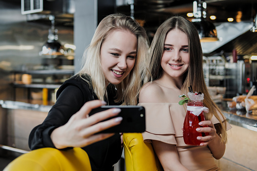 Two young smart beautiful girls in dresses are sitting at the bar. Drink red and blue cocktails from glasses with tubules, communicate and laugh. Take a selfie on the phone.