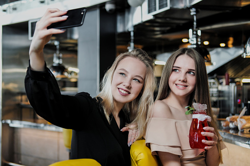 Two young smart beautiful girls in dresses are sitting at the bar. Drink red and blue cocktails from glasses with tubules, communicate and laugh. Take a selfie on the phone.