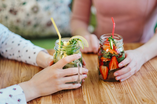 Close-up of the hands of two girls holding glass jars with red strawberry and yellow lemon lemonade. The concept of summer soft drinks.