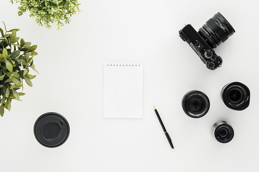 Mocap work photographer on a white table. Flat lay mirrorless camera, a few lenses and a notebook with a pen. Green pot plants. Minimalism.