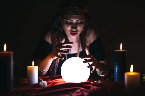 Young female fortune teller is looking into glowing crystal sphere