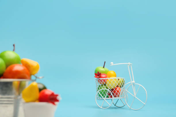 bicicleta de alambre blanco con una cesta llena de mini fruta. cerca del cubo con verduras y frutas. fondo azul. concepto de recolección y entrega de frutas y hortalizas. - abundance apple red yellow fotografías e imágenes de stock