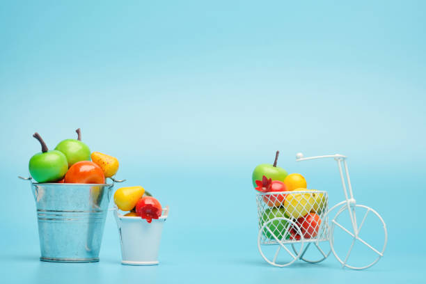 bicicleta de alambre blanco con una cesta llena de mini fruta. cerca del cubo con verduras y frutas. fondo azul. concepto de recolección y entrega de frutas y hortalizas. - abundance apple red yellow fotografías e imágenes de stock