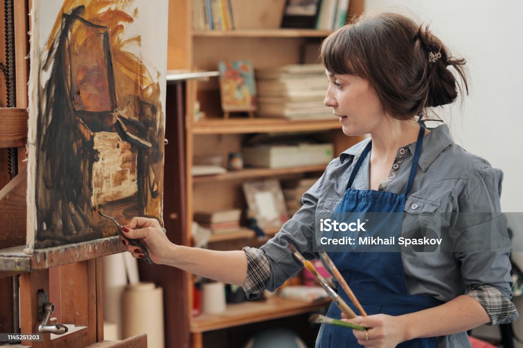 Young beautiful woman painter among easels and canvases in a bright studio. Young beautiful woman painter among easels and canvases in a bright studio. Inspiration and hobby. Painting - Activity Stock Photo