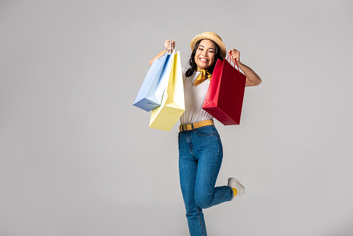 Happy trendy dressed asian woman holding colorful shopping bags on raised hands isolated on grey