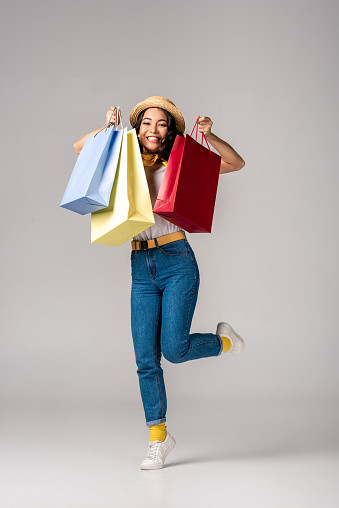 Happy trendy dressed asian woman holding colorful shopping bags on raised hand on grey