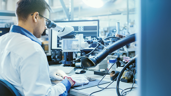 El trabajador de fábrica de electrónica en el abrigo de trabajo blanco inspecciona una placa de circuito impreso a través de un microscopio digital. Instalaciones de fábrica de alta tecnología. photo