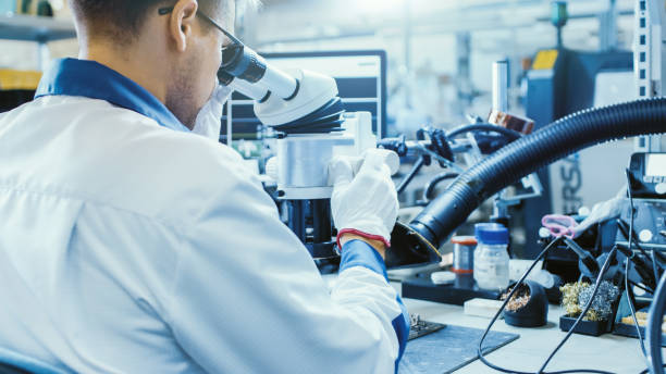 istock Electronics Factory Worker in White Work Coat is Soldering a Printed Circuit Board Through a Digital Microscope. High Tech Factory Facility. 1145276194