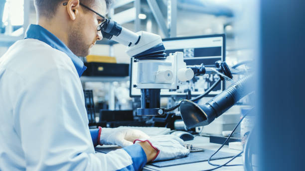 istock Electronics Factory Worker in White Work Coat is Soldering a Printed Circuit Board Through a Digital Microscope. High Tech Factory Facility. 1145276205