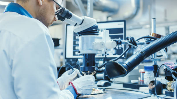 istock Electronics Factory Worker in White Work Coat is Soldering a Printed Circuit Board Through a Digital Microscope. High Tech Factory Facility. 1145276211