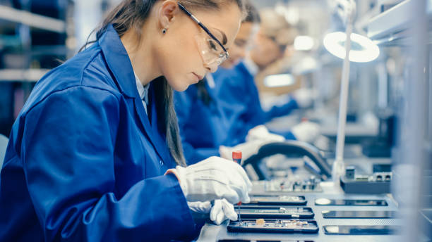 istock Female Electronics Factory Workers in Blue Work Coat and Protective Glasses Assembling Printed Circuit Boards for Smartphones with Tweezers. High Tech Factory with more Employees in the Background. 1145276246