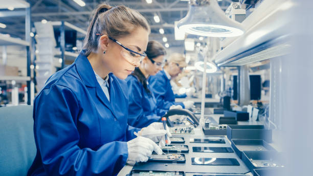 istock Woman Electronics Factory Worker in Blue Work Coat and Protective Glasses is Assembling Smartphones with Screwdriver. High Tech Factory Facility with more Employees in the Background. 1145276247