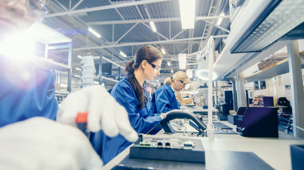 istock Female Electronics Factory Worker in Blue Work Coat and Protective Glasses is Assembling Laptop's Motherboard with a Screwdriver. High Tech Factory Facility with Multiple Employees. 1145276249