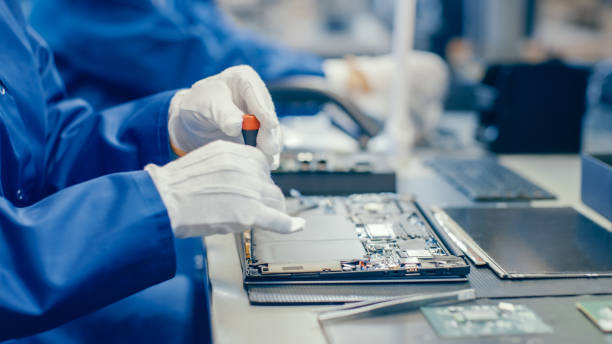 istock Close-Up of a Female Electronics Factory Worker in Blue Work Coat Assembling Laptop's Motherboard with a Screwdriver. High Tech Factory Facility with Multiple Employees. 1145276264