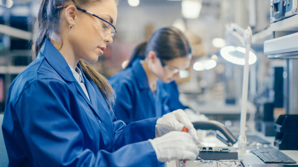 istock Female Electronics Factory Worker in Blue Work Coat and Protective Glasses is Assembling Laptop's Motherboard with a Screwdriver. High Tech Factory Facility with Multiple Employees. 1145276265