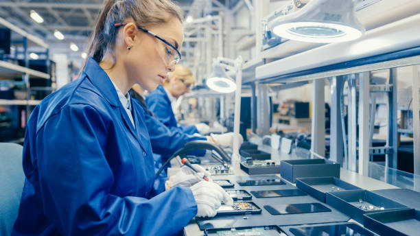 istock Woman Electronics Factory Worker in Blue Work Coat and Protective Glasses is Assembling Smartphones with Screwdriver. High Tech Factory Facility with more Employees in the Background. 1145276294
