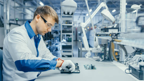istock Young Man in Blue and White Work Coat is Using Plier to Assemble Printed Circuit Board for Smartphone. Electronics Factory Workers in a High Tech Factory Facility. 1145276300