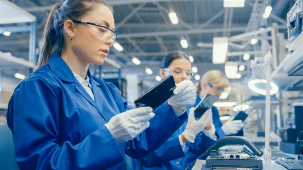 istock Female Electronics Factory Worker in Blue Work Coat and Protective Glasses Takes a Smartphone Screen and Performs a Quality Check. High Tech Factory Facility with Multiple Employees. 1145276302