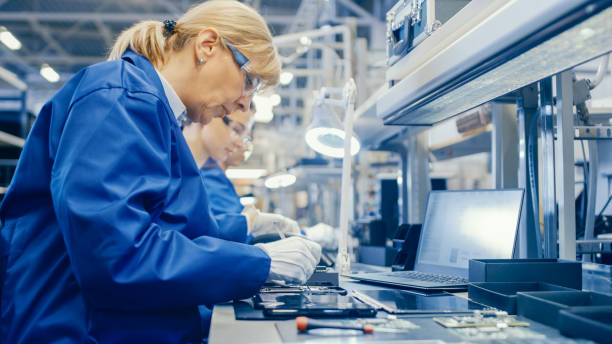 istock Female Electronics Factory Worker in Blue Work Coat and Protective Glasses is Assembling Smartphones with Screwdriver. High Tech Factory Facility with more Employees in the Background. 1145276307