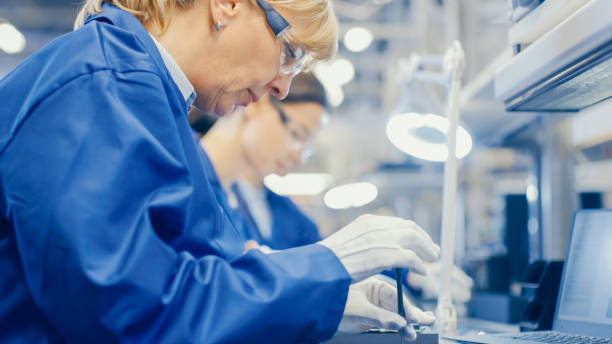 istock Female Electronics Factory Worker in Blue Work Coat and Protective Glasses is Assembling Smartphones with Screwdriver. High Tech Factory Facility with more Employees in the Background. 1145276319