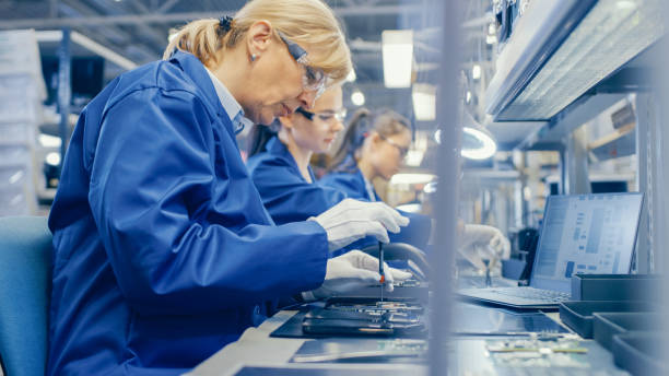 istock Female Electronics Factory Worker in Blue Work Coat and Protective Glasses is Assembling Smartphones with Screwdriver. High Tech Factory Facility with more Employees in the Background. 1145276336