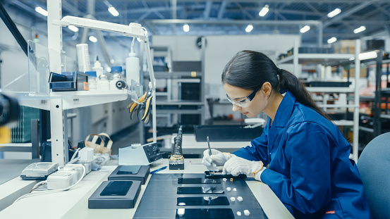 Joven mujer en el abrigo de trabajo azul es ensamblar placas de circuito impreso para smartphones. Trabajadores de la fábrica de electrónica en una fábrica de alta tecnología. photo