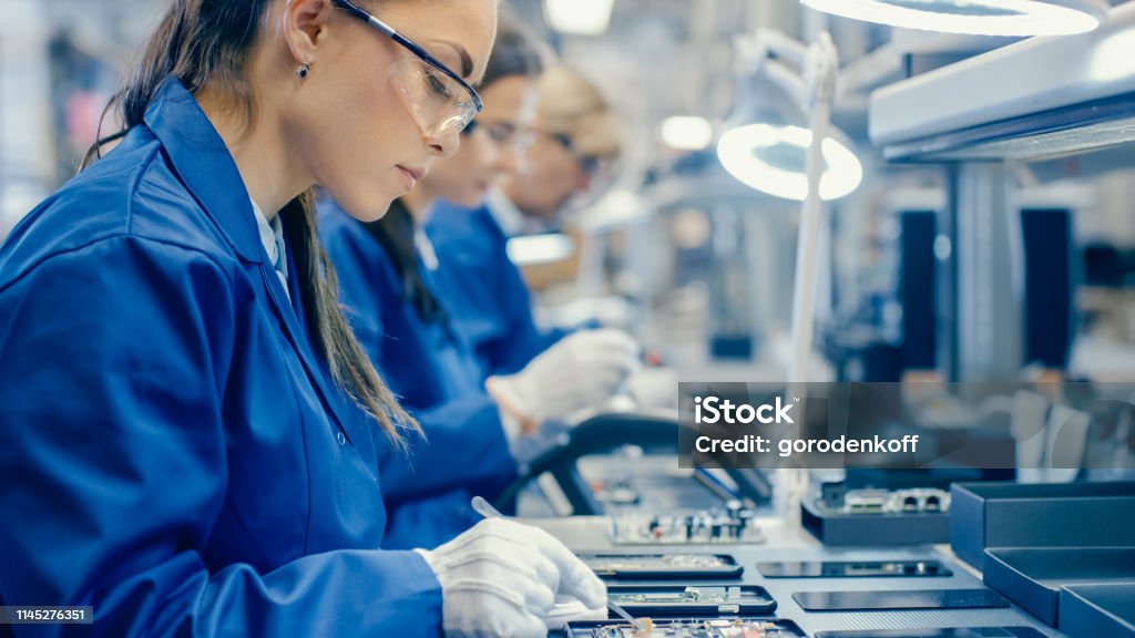 Female Electronics Factory Workers in Blue Work Coat and Protective Glasses Assembling Printed Circuit Boards for Smartphones with Tweezers. High Tech Factory with more Employees in the Background. Manufacturing Stock Photo