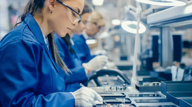 Female Electronics Factory Workers in Blue Work Coat and Protective Glasses Assembling Printed Circuit Boards for Smartphones with Tweezers. High Tech Factory with more Employees in the Background.