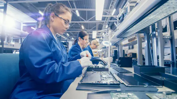 Woman Electronics Factory Worker in Blue Work Coat and Protective Glasses is Assembling Laptop's Motherboard with a Screwdriver. High Tech Factory Facility with Multiple Employees.