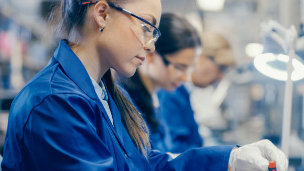istock Female Electronics Factory Worker in Blue Work Coat and Protective Glasses is Assembling Laptop's Motherboard with a Screwdriver. High Tech Factory Facility with Multiple Employees. 1145276373